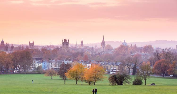 Eine Panoramaansicht einer Stadt mit einer Skyline aus Türmen und grünen Feldern im Vordergrund bei Sonnenuntergang.