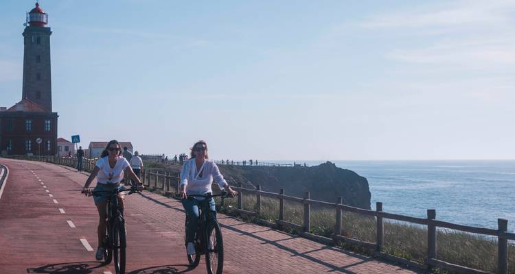 Dos mujeres andando en bicicleta por una carretera costera junto a un faro en una mañana despejada.