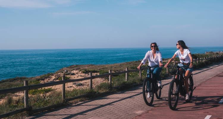 Dos ciclistas andan en bici lado a lado junto al océano en un camino pavimentado con dunas ondulantes y agua azul a su lado.
