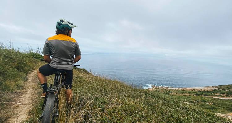 Un ciclista de montaña descansa en un sendero de acantilado cubierto de pasto mirando hacia un horizonte oceánico infinito.