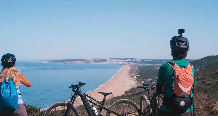 Los ciclistas se detienen en un sendero en lo alto del acantilado admirando una larga playa de arena y el pueblo costero muy abajo.