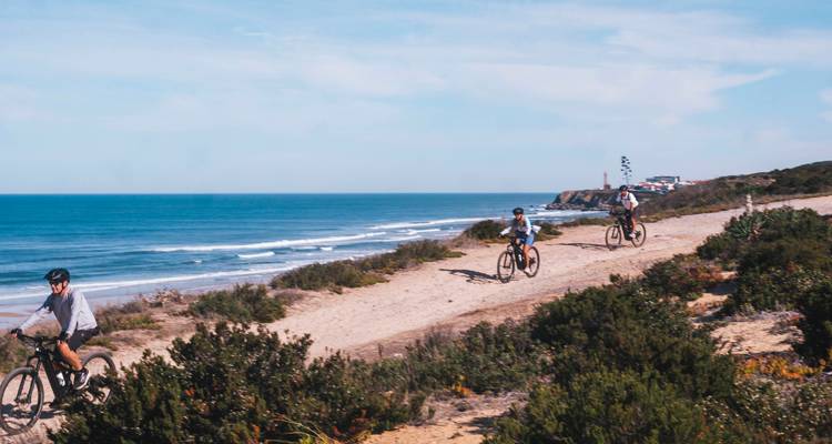 Tres ciclistas de montaña conducen por un sendero costero arenoso junto a las olas del océano turquesas.