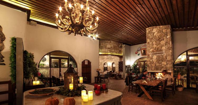 Sala de comedor rústica con iluminación cálida, paredes de piedra, lámpara de araña e invitados cerca de una chimenea.
