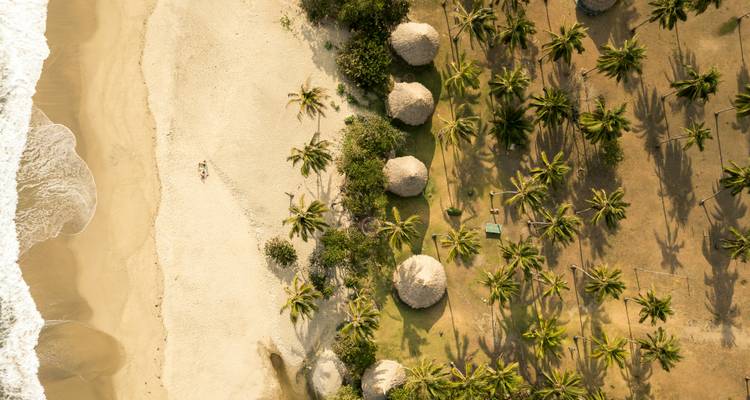 Foto aérea con dron de una playa caribeña bordeada de palmeras con arena blanca, cabañas con techo de paja y olas turquesas.