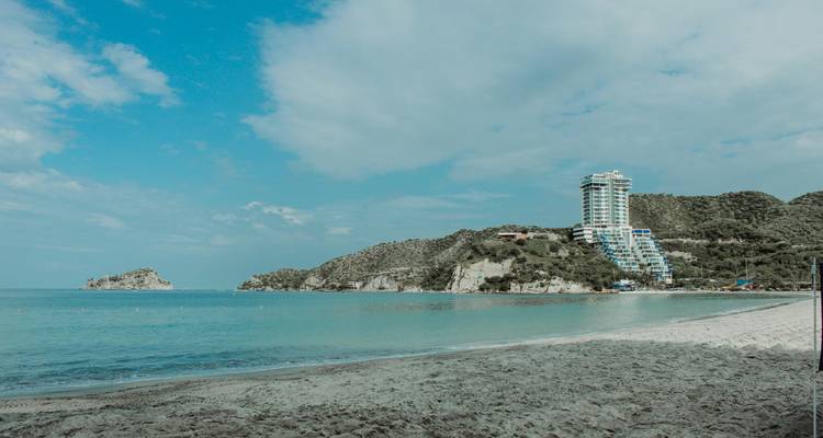 Playa tranquila frente a la ciudad con arena suave, agua cristalina y un moderno hotel de gran altura rodeado de colinas verdes.
