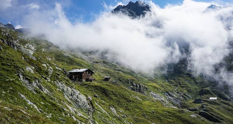 Une cabane de montagne isolée se dresse sur une pente verte voilée de brume avec des sommets imposants partiellement cachés par les nuages.