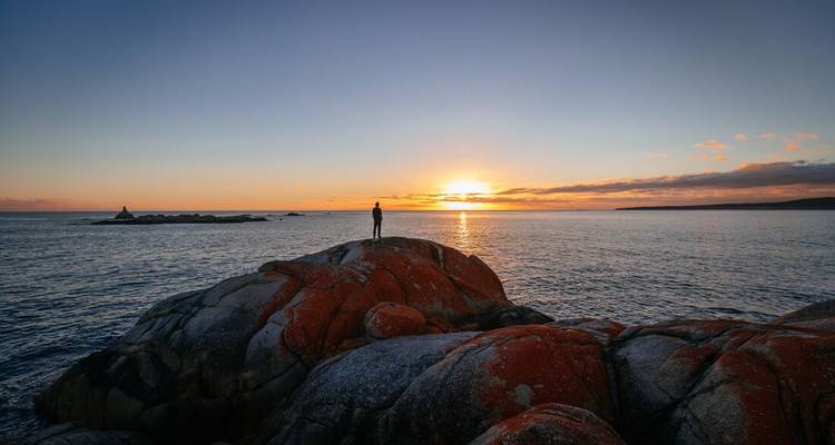 Silhouette solitaire se tenant sur des rochers recouverts de lichen orange observant le coucher de soleil sur la mer