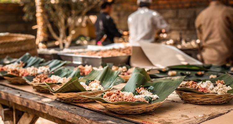 Nourriture balinaise traditionnelle servie sur des feuilles de bananier avec des personnes préparant de la nourriture en arrière-plan.