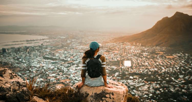 Personne assise sur un rocher contemplant un vaste paysage urbain au coucher du soleil.