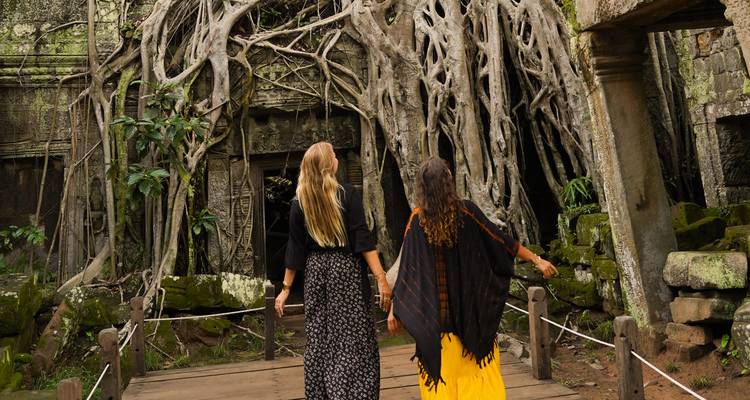 Zwei Frauen erkunden einen antiken Tempel, der von großen Wurzeln überwuchert ist.
