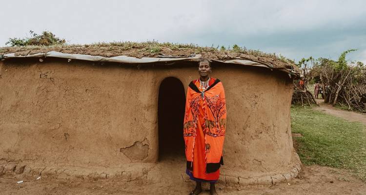 Une femme Maasai vêtue d'un shuka rouge vif se tenant devant une hutte traditionnelle en terre.