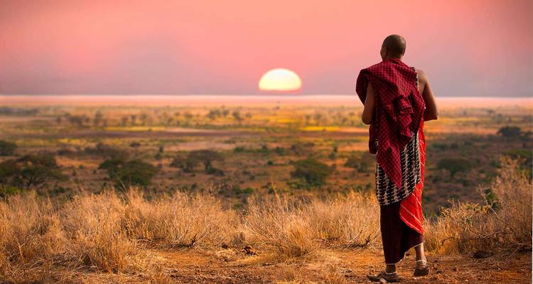Un homme maasaï vêtu de ses habits traditionnels contemple un coucher de soleil dramatique de couleur orange au-dessus de la savane africaine.