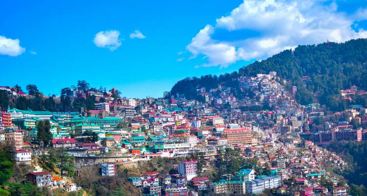 Kleurrijke heuvelachtige stadsgezicht van Shimla onder een helder blauwe lucht en wollige wolken.