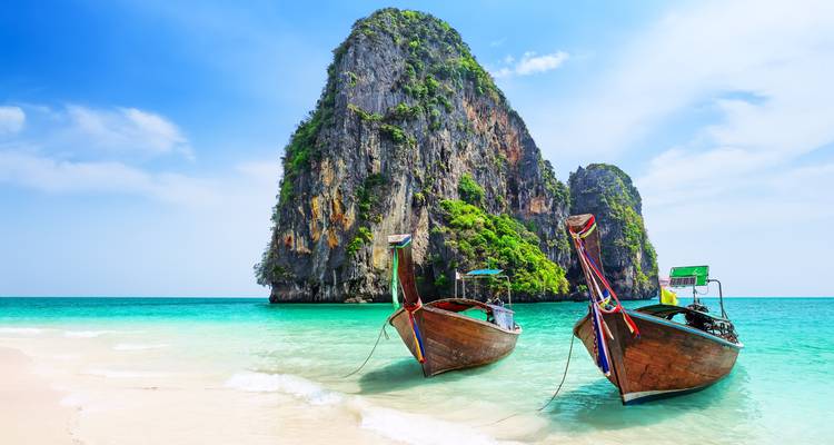 Traditional Thai boats on a clear sea with a limestone cliff in the background.