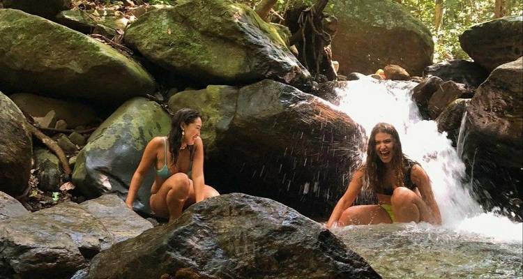 Two women sitting on rocks near a small waterfall in a lush green environment.