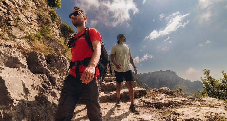 Deux hommes faisant de la randonnée sur un sentier rocheux avec un arrière-plan montagneux et un ciel partiellement nuageux.