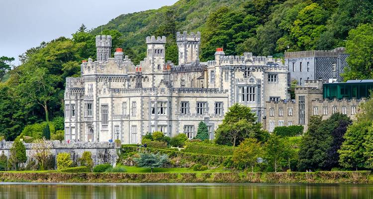A grand historic castle, nestled in the greenery by a lake, with a reflection in the water.