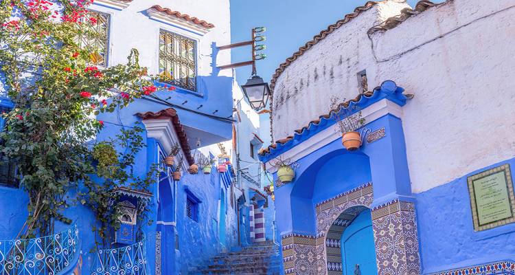 Ruelles bleues vibrantes de Chefchaouen ornées de carreaux, de fleurs et de pots décoratifs sous un ciel dégagé.
