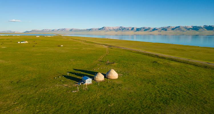 Luftaufnahme von zwei Jurten auf der grünen Steppe neben dem schimmernden Song-Kul-See und einer fernen Bergkette.