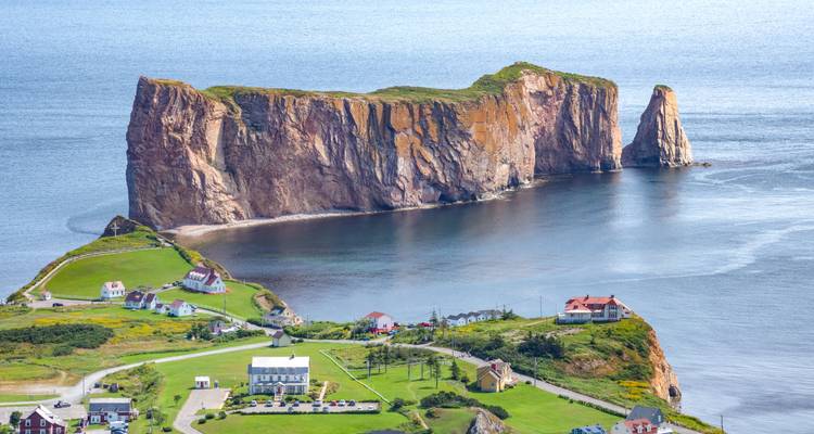 Luchtfoto van Percé Rock die dramatisch oprijst uit de zee naast een klein kustdorpje met groene gazons en huizen.