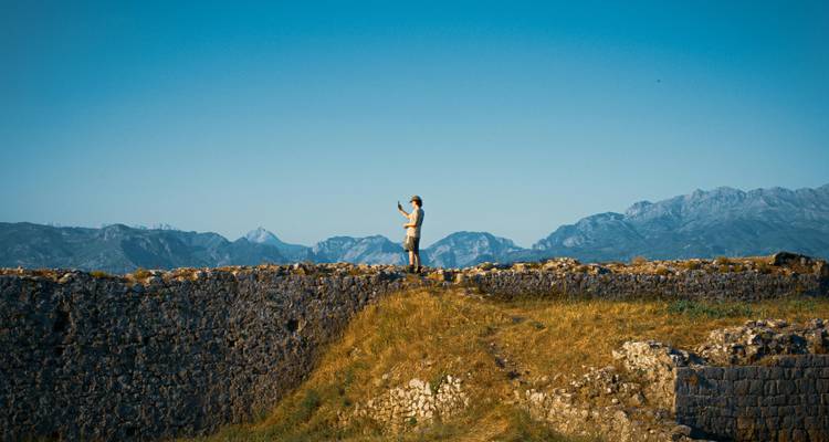 Person standing on a stone wall with mountains in the distance.
