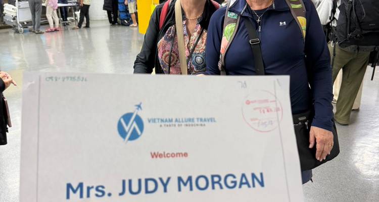 Viajeros que llegan posando en un aeropuerto mientras se sostiene un letrero que dice 'Mrs. Judy Morgan' frente a ellos.