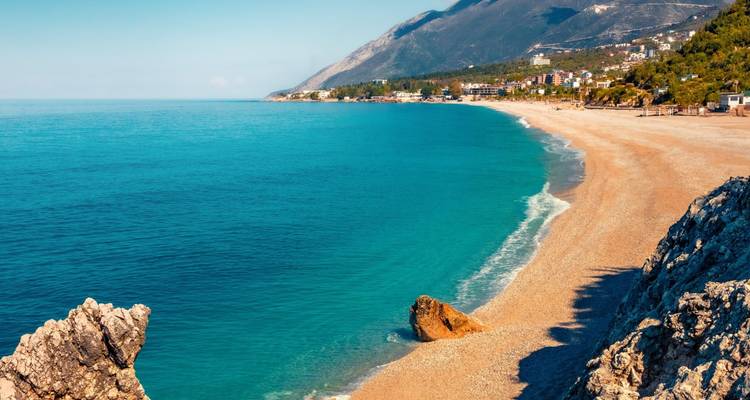 Plage de sable blanc étendue avec une eau turquoise et des montagnes s'élevant derrière un village côtier situé le long de la Riviera albanaise.