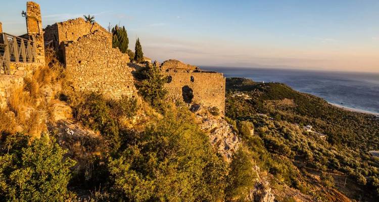 Lumière de l'heure dorée sur les murs de pierre anciens d'une ruine perchée sur une colline dominant des pentes couvertes d'oliviers et la mer.