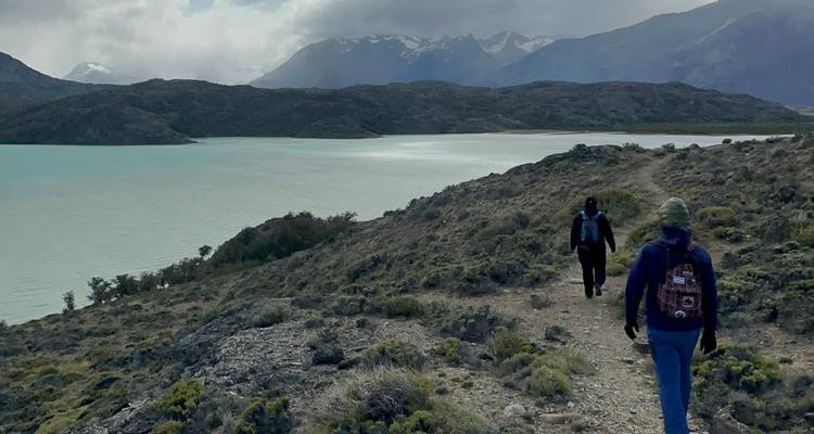 Dos excursionistas caminando por un sendero junto al lago con montañas.