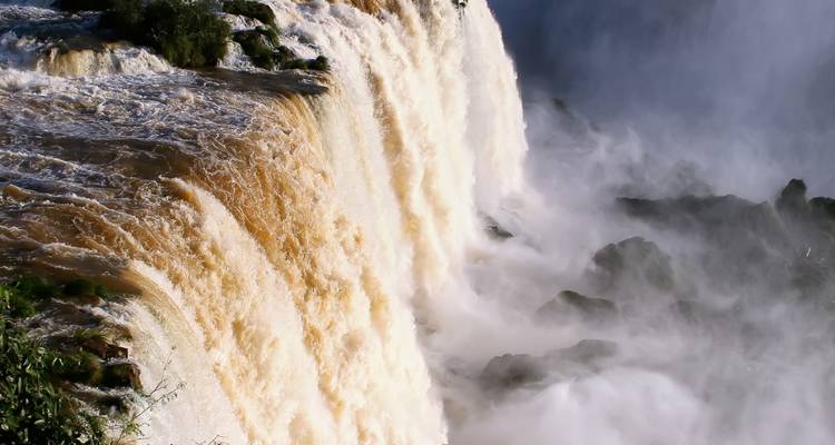 Cascada poderosa con neblina y rocas, arcoíris visible.