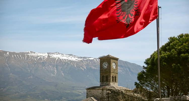 Le drapeau albanais flotte au-dessus d'une tour d'horloge en pierre sur un mur de forteresse avec des montagnes enneigées en arrière-plan.