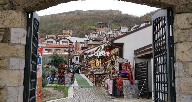 Calle de mercado bulliciosa enmarcada por una puerta de piedra, bordeada de puestos de artesanía coloridos y fortaleza en la ladera al fondo.