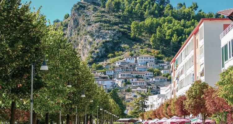 Boulevard soleado con sombrillas de cafés que lleva a una ladera repleta de casas otomanas blancas tradicionales y frondosos árboles verdes.