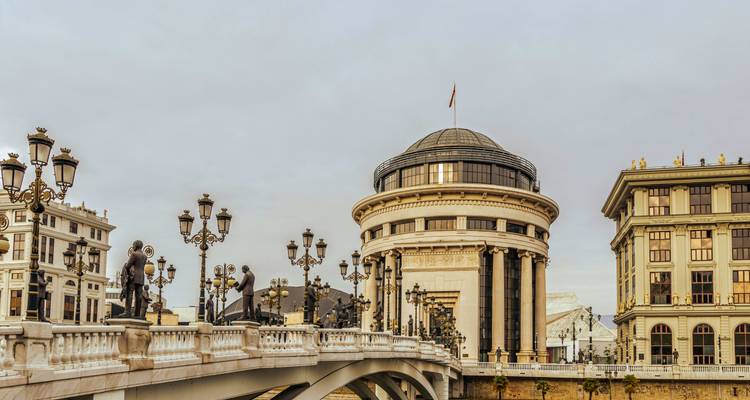 Un elegante puente de piedra con estatuas y farolas ornamentadas que lleva a un edificio de museo neoclásico con cúpula en un paisaje urbano.