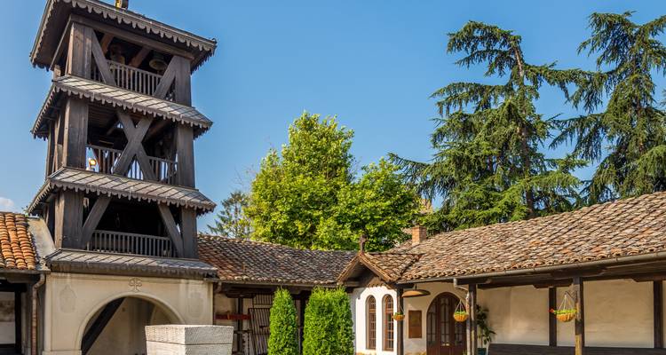 Torre de campanas de madera que se eleva sobre un apacible patio con tejados de azulejos, paredes encaladas de blanco y altos árboles de hoja perenne.