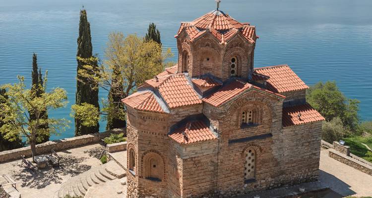 Pintoresca iglesia bizantina de San Juan en Kaneo a orillas del lago, encaramada en un acantilado sobre aguas azul profundo y rodeada de cipreses.