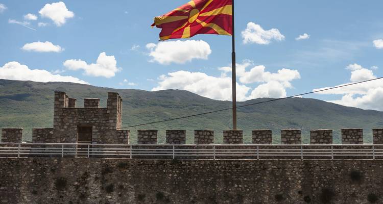 La bandera macedónica ondeando sobre la pared de piedra de una fortaleza con montañas y cielo azul de fondo.