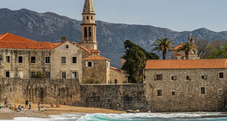Complexe monastique aux murs de pierre et clocher sur le littoral adriatique de Budva avec des montagnes s'élevant en arrière-plan.