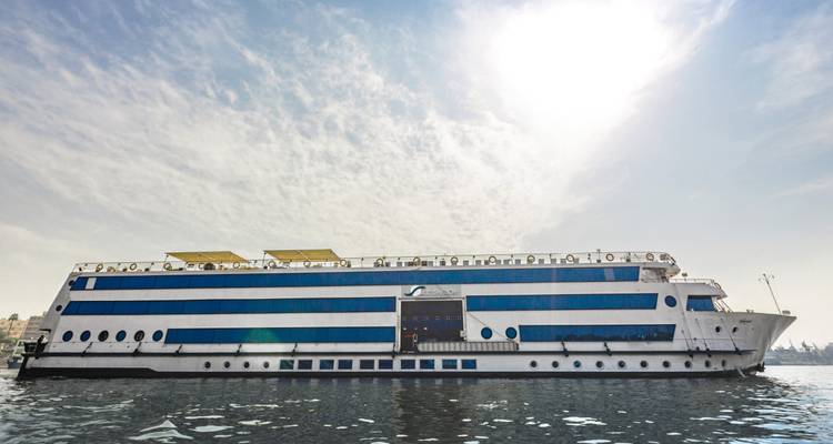 Grand navire de croisière blanc et bleu sur le Nil naviguant sur des eaux calmes sous un soleil de midi éclatant et des nuages légers.