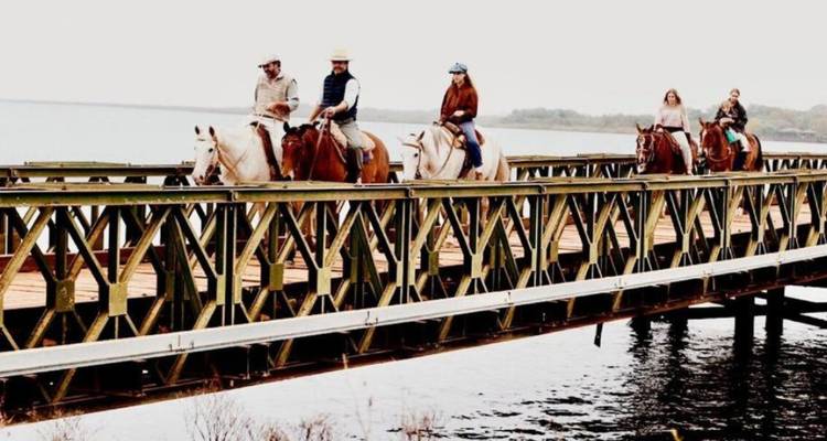 Grupo de personas montando caballos en un puente sobre el agua.