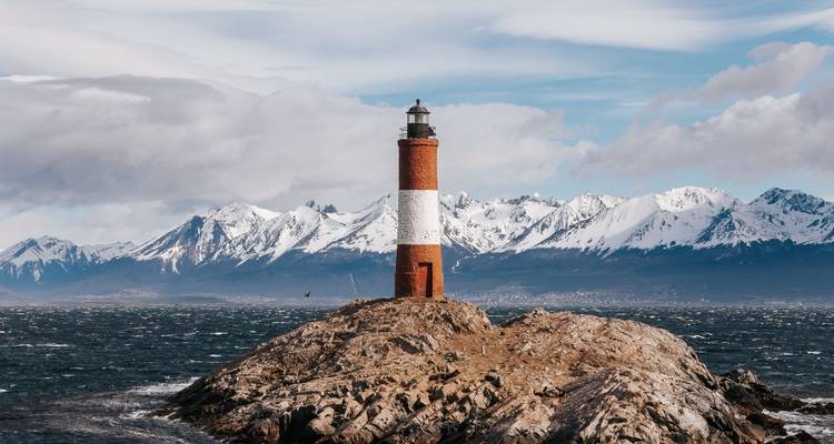 Phare sur une île rocheuse avec des montagnes enneigées en arrière-plan.