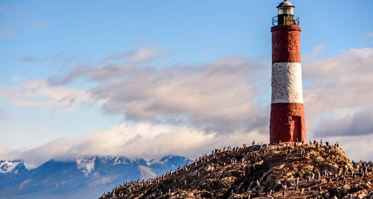 Phare sur une île rocheuse grouillant d'oiseaux, avec des montagnes en arrière-plan.
