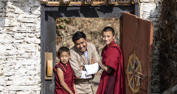Ein Mann und zwei junge Mönche lächeln und stehen neben einem kunstvoll gestalteten Tor.