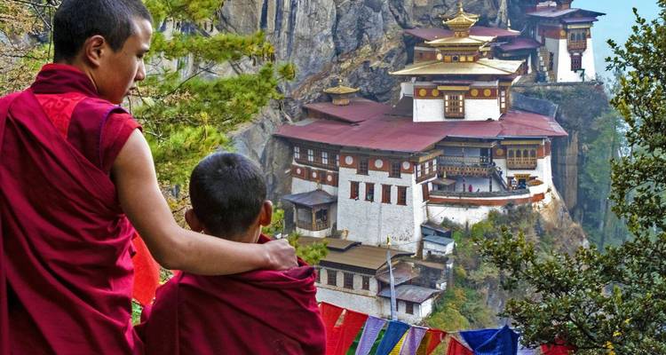 Zwei Mönche stehen auf einer Klippe und blicken auf das Tigernest-Kloster mit Gebetsfahnen.