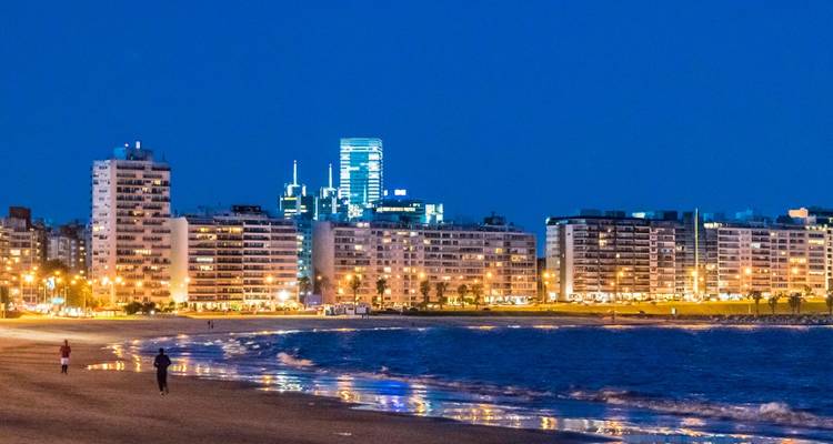 Nachtgezicht van een strand met stadslichten en reflecties op het water.