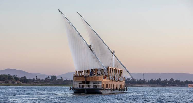 Un elegante dahabiya de dos mástiles de madera navega serenamente por el Nilo al atardecer.