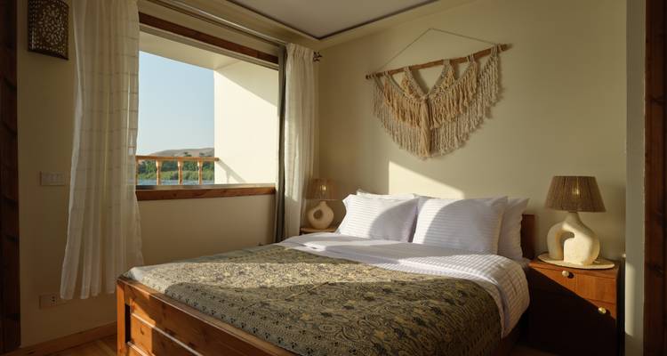 Acogedor dormitorio de cabaña con decoración tejida y luz solar entrando a través de una ventana con vistas al Nilo.