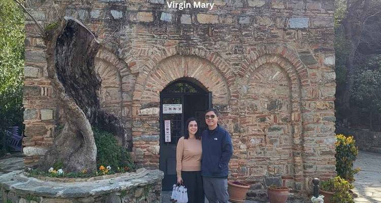 Una pareja se encuentra frente a la casa de piedra de la Virgen María con texto descriptivo en la parte superior de la foto.