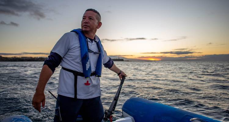Man navigating a boat at sunset with water in the background.