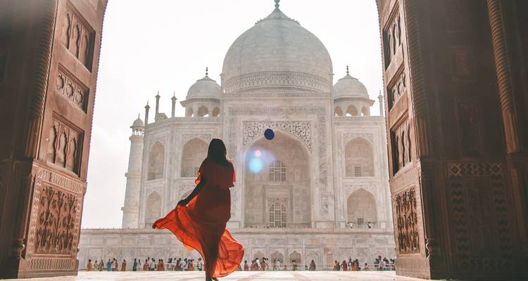 A woman in a flowing red dress looking at the Taj Mahal through an archway.